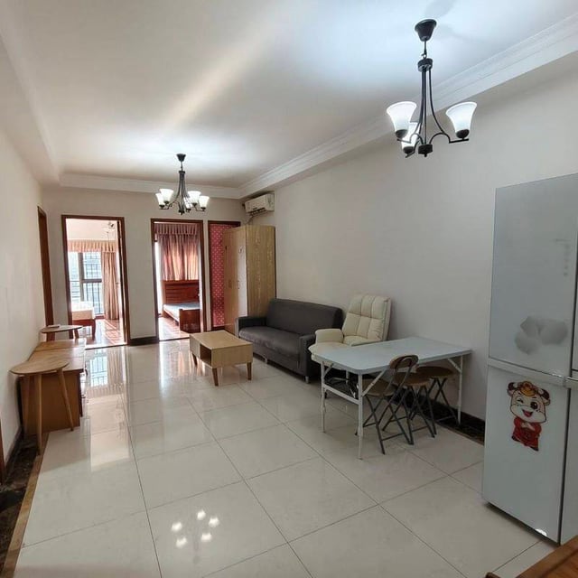 Living room with a grey sofa, desk, refrigerator and open bedroom door in KONGS's Xiangnan Ruifeng Garden listing photo